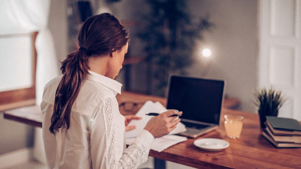 部屋の中の背景。女性がノートパソコンが置かれたテーブルで資料を読み書きしている画像。