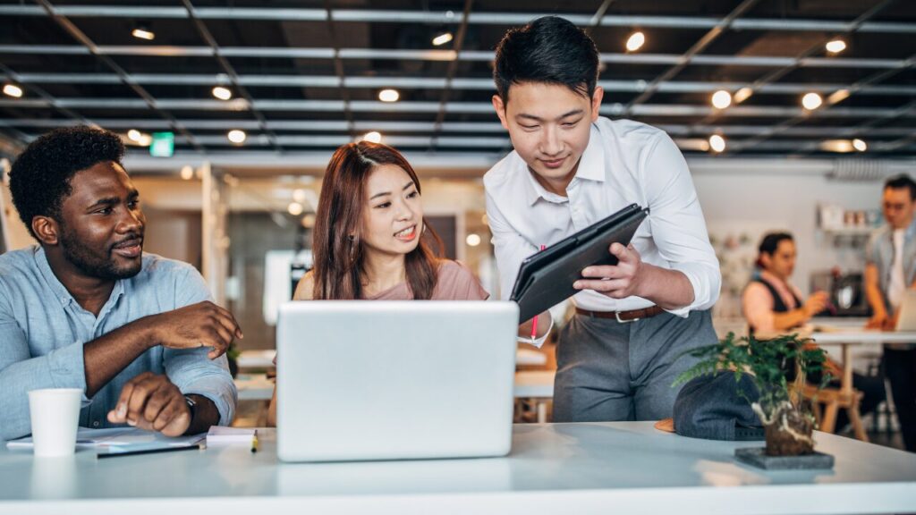 コワーキングスペースで3人の人物がタブレットやPCを見ながら会話をしている画像